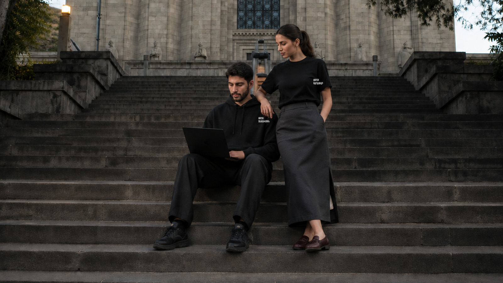 Two models on stone steps in Yerevan wearing a black URISH hoodie and T-shirt beside a laptop